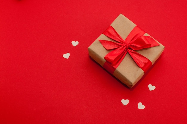 Gift box in craft packaging and with a red ribbon on a red background with hearts