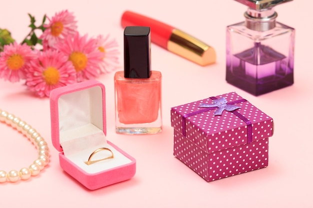 Подарочная коробка, цветы, флаконы с лаком для ногтей и парфюмерия, помада, золотое кольцо в коробке, бусины на розовом фоне. Женская косметика и аксессуары.