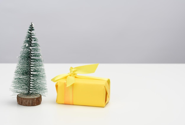 Gift box is wrapped in yellow paper and a decorative Christmas tree on a white table. Festive background