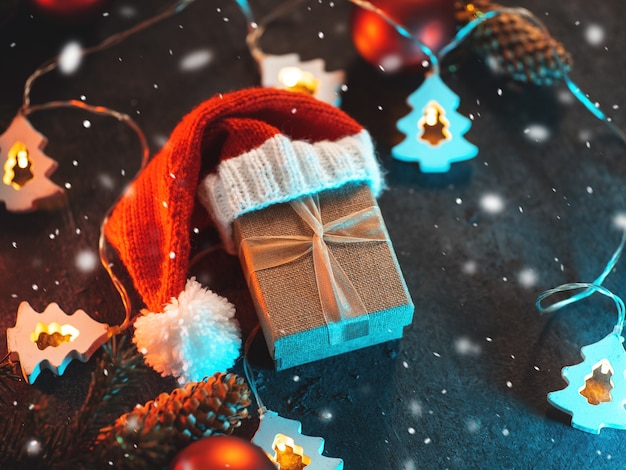 A gift box in a knitted Santa hat on a dark table with a garland, branches of a Christmas tree with cones. Christmas background