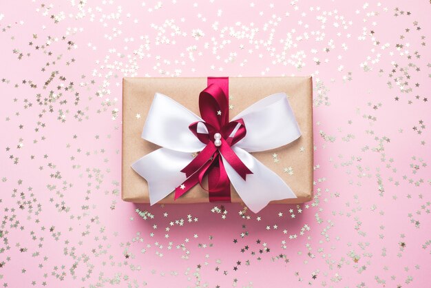 Gift box on a pink background with gold confetti stars. Present in a box in a bow.