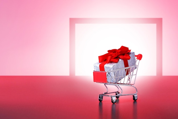 Gift box in shopping cart near supermarket door on pink background.