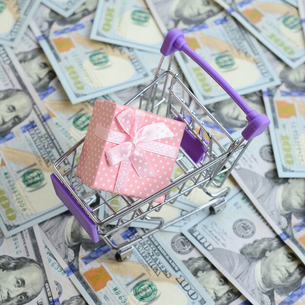 Gift box in a small shopping cart lies on a many dollar bills