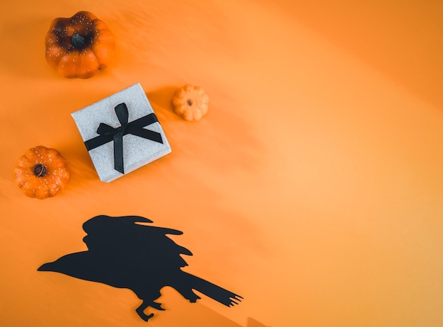 Gift box three pumpkins and a paper witch on a raven lie on the left against an orange background with space for text on the right closeup flat lay