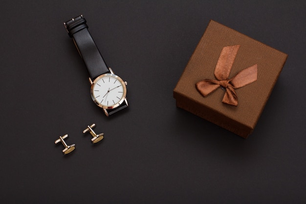 Gift box, watch with a black leather strap and cufflinks on a black background. Accessories for men. Top view.