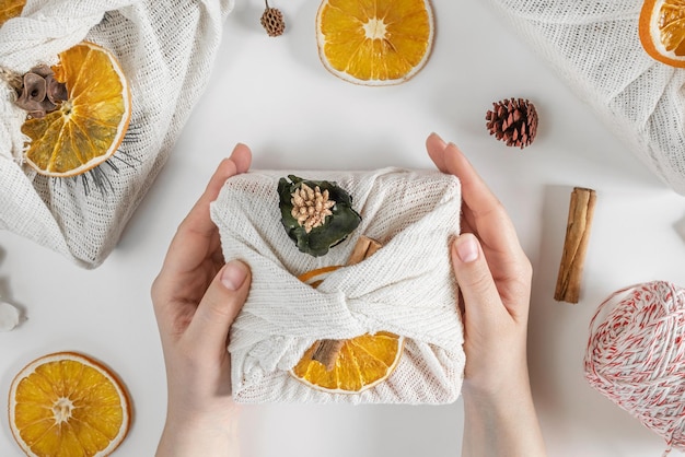 Gift Box in White Fabric. Female Hands wrapping Sustainable Gift on white Table with Decoration Elem