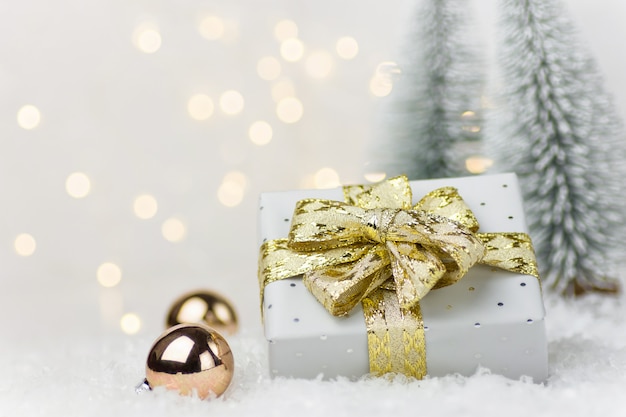 Gift box with golden ribbon bow baubles in winter forest with fir trees snow