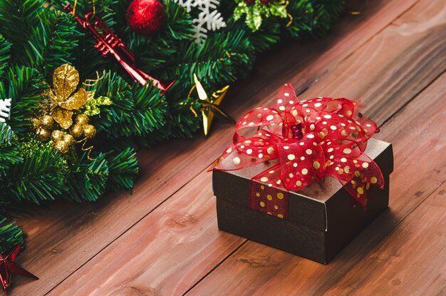 Gift box with red color ribbon on wood background with Christmas tree and traditional ornaments Xmas props with copy space. Concept of happy and joyful in festival.