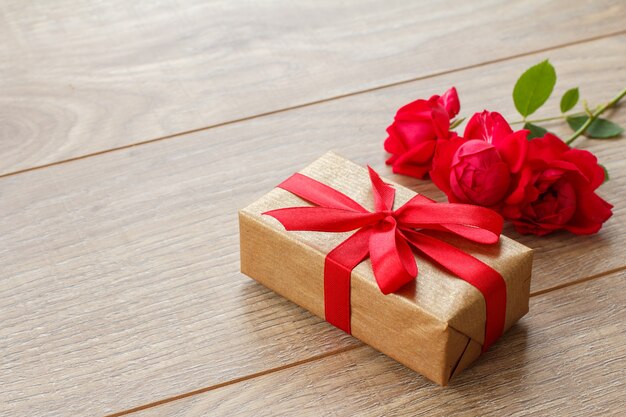 Gift box with red ribbon on wooden boards with bouquet of red roses. Top view with copy space.