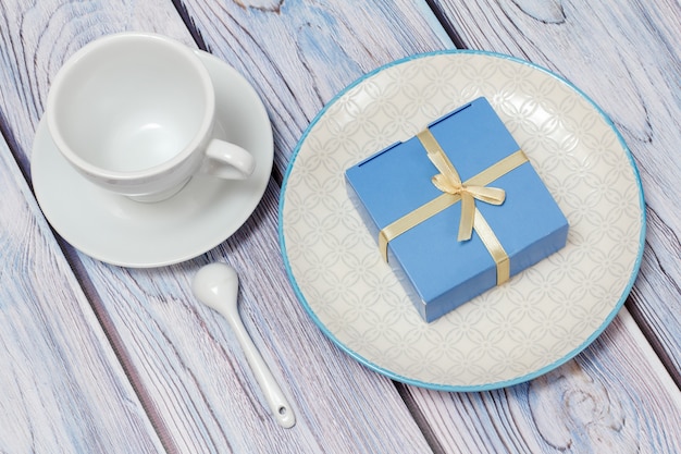 Gift box with ribbons on the white plate and a cup with saucer on the wooden boards