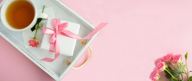 분홍색 배경의 흰색 나무 쟁반에 있는 컵에 묶인 분홍색 활과 차가 있는 선물 상자Top view Copy space