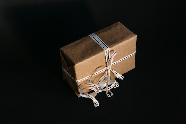 Gift box wrapped in recycled craft paper and tied with a white and gold ribbon bow on a black background. Surprise for the holiday.