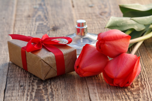 Gift box wrapped with red ribbon, red tulips and bottle of perfume on wooden boards.