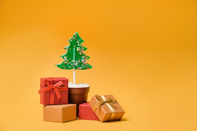 Gift boxes near the Christmas tree, on a yellow background with copy space. Lollipop in the form of a Christmas tree in a flower pot with Christmas gifts.