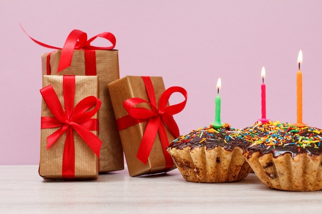 ピンクの背景に燃えるお祝いのキャンドルで飾られたギフトボックスとチョコレート釉薬とキャラメルの3つのおいしい誕生日カップケーキ。お誕生日おめでとうミニマルコンセプト。