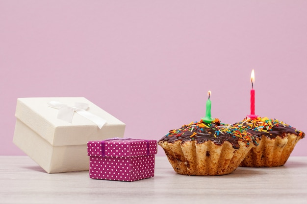 Gift boxes and two tasty birthday muffins with chocolate glaze and caramel, decorated with burning festive candles on lilac background. Happy birthday minimal concept.