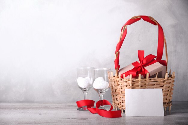 Gift boxes with red ribbons on a basket, two white hearts in wine glasses and blank love letter