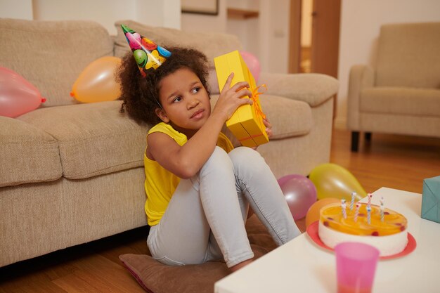 Gift. a dark-skinned girl in a bday hat sitting on the floor and holding a gift