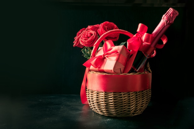 Подарочная корзина, букет красных роз, бутылка шампанского на черном фоне.