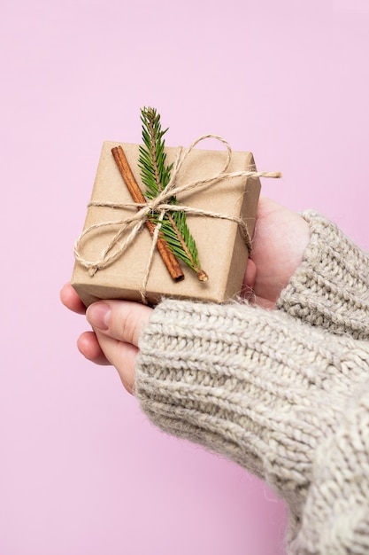 Gift made of Kraft paper and a Christmas tree branch in women's hands on a pink background, close-up. Handmade gifts concept. Modern trend, natural gift wrapping with your own hands.