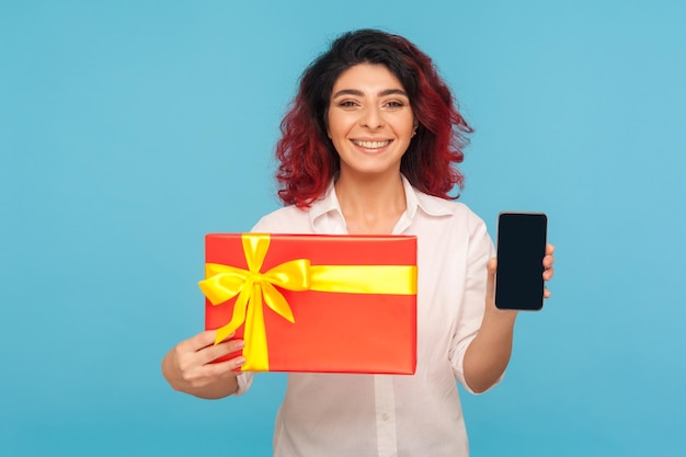 Подарочный заказ онлайн Портрет счастливой улыбающейся женщины с причудливыми рыжими волосами, показывающей завернутую коробку и мобильный телефон, выглядящей довольной настоящим мобильным сервисом в помещении студии, снятой на синем фоне