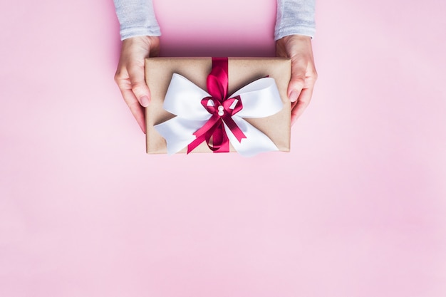 Gift or present box with a big bow in the hands of a woman on a pink table. Flat lay composition for Christmas, birthday, mother day or wedding.