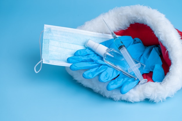 Gift set for Christmas and New Years 2021 holidays. A hygienic set of a medical mask, gloves, a disinfectant and a syringe with a vaccine and a coronavirus chip in a Santa Claus hat. Vaccination 2021.