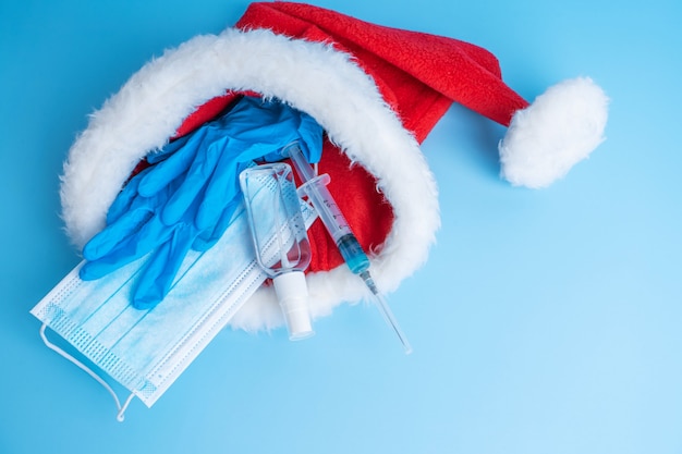 Gift set for Christmas and New Years 2021 holidays. A hygienic set of a medical mask, gloves, a disinfectant and a syringe with a vaccine and a coronavirus chip in a Santa Claus hat. Vaccination 2021.