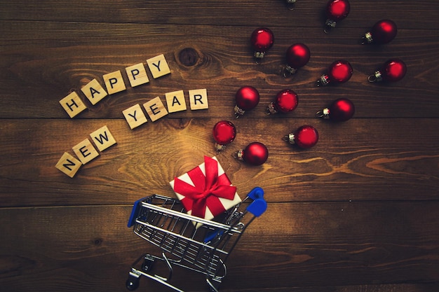 Подарок в маленькой тележке, надпись деревянными буквами с новым годом и разноцветные елочные шары на деревянном фоне. Темный новогодний фон.