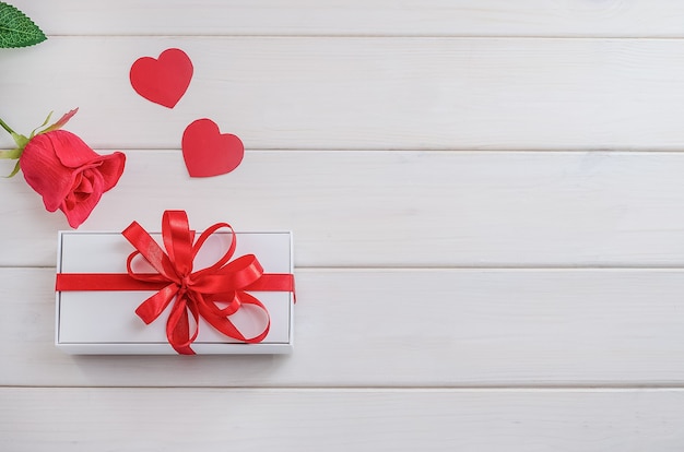 Gift for Valentine's Day, Mother's Day. White gift box with a red bow with a rose with hearts with copy space on a white wooden background. Holiday Sale, Congratulations