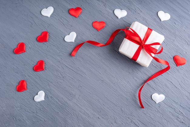 Подарок в белой бумаге с красной лентой, бело-красной формой сердца на стильном сером гранж-фоне