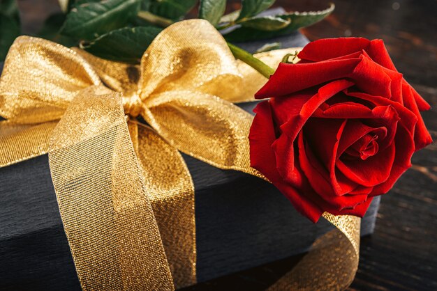 Gift wrapped in black paper and a gold ribbon. on top of the box is a luxurious red rose.