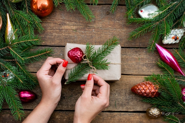 Gift wrapped in craft paper from a sprig of spruce. Decor from Christmas tree decorations and branches of a Christmas tree. Hands untie the gift.