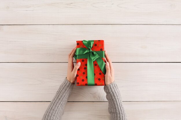 Gift wrapping background. Female hands holding handmade present box in red dotted paper against white wooden table background. Bithday or christmas preparation concept, top view, copy space