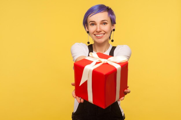 Gift for you. Portrait of cute happy trendy girl with violet short hair in overalls giving big red present box to camera and smiling, charity concept. isolated on yellow background, studio shot