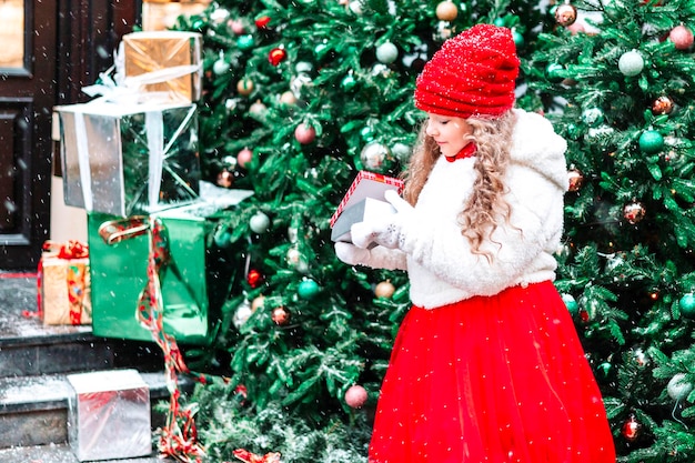 クリスマスと新年の贈り物 クリスマス ツリーの隣に白い毛皮のコートと赤い帽子とスカートを着た女の子が屋外のお祝いと贈り物を受け取ります