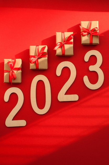 Gifts in craft packaging with red ribbons and the number 2023 on a red background with shadows with space for text in vertical format