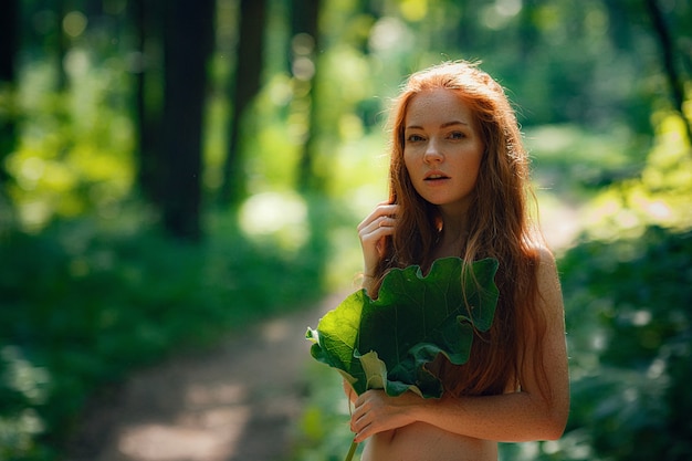 Ginger beautiful woman holding a big leave over her chest with no clothes under in a forest
