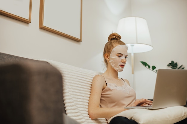 Ginger caucasian lady wearing an anti aging mask while relaxing on the sofa with a computer