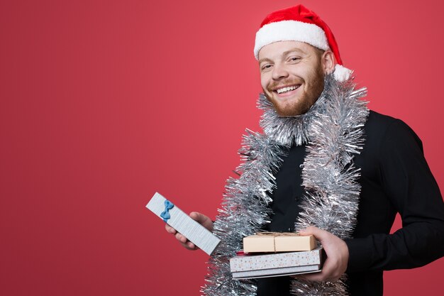 Ginger man with santa hat is advertising something while holding new year present on red  wall