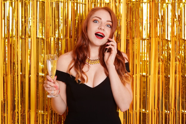 Ginger shocked young woman in little black dress holding glass of champagne