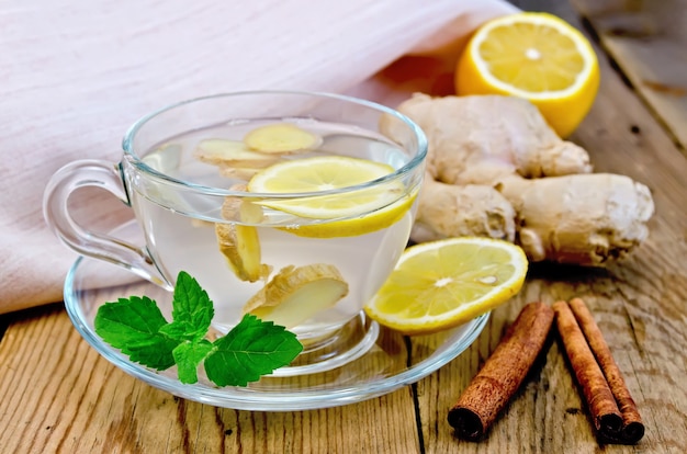 Ginger tea in a glass cup, lemon, cinnamon, ginger, mint, napkin against a wooden board