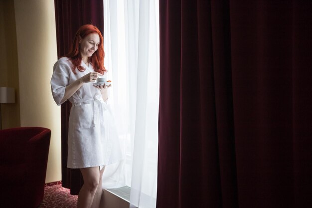 A ginger woman standing by the window and holding a cup of coffee looking at the cup
