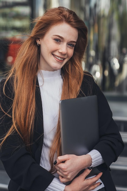 Foto ginger zakenvrouw poseren buiten in jas met laptop voor werk kijken en glimlachen naar de camera het verlaten van kantoor het is tijd voor een pauze