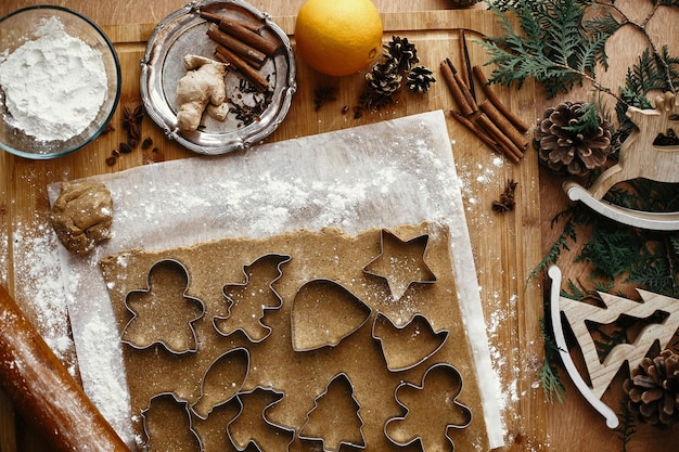 Gingerbread dough with metal cutters in different shapes for christmas cookies and wooden rolling pin anise ginger cinnamon pine cones fir branches flour on rustic table Flat lay