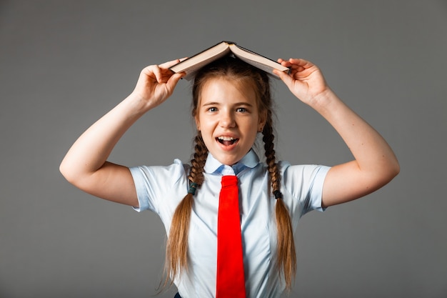 Girl 12 years old in school uniform with book above head isolated on gray background