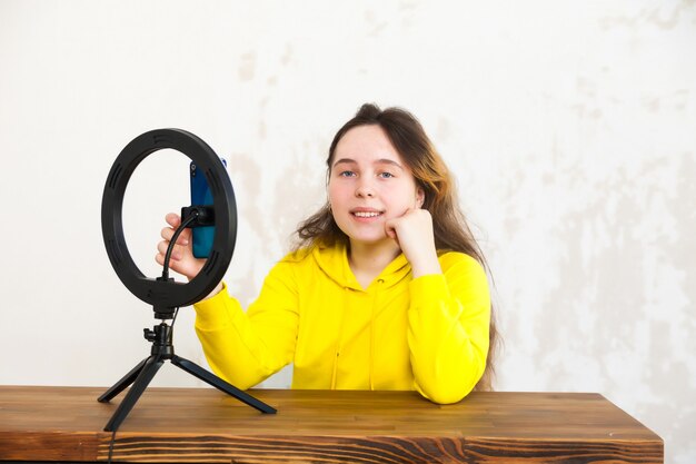 Girl of 16 years old recording video on smartphone and lighting herself with ring lamp at table in light room