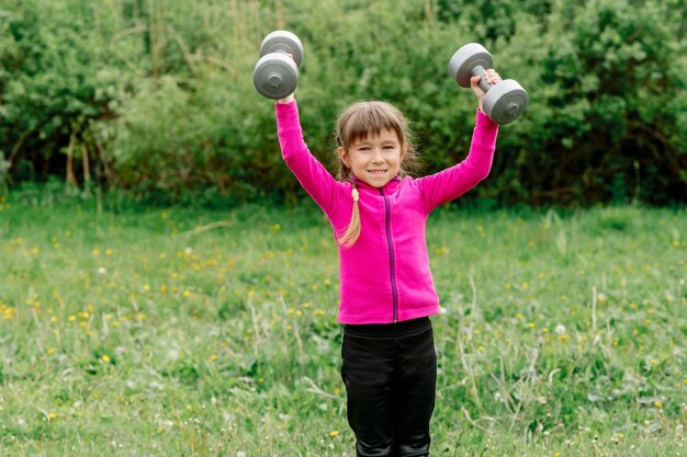 A girl 5 years old in a tracksuit raises dumbbells over her head in nature