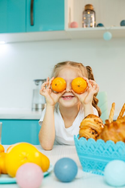 A girl of 7-8 years old sits in the kitchen and makes eyes with a tangerine.