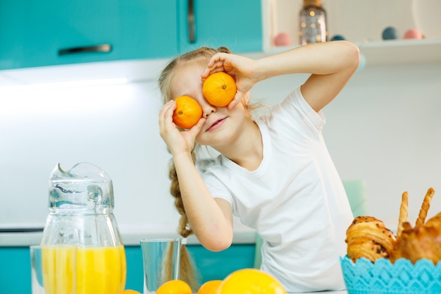 a girl of 7-8 years old sits in the kitchen and makes eyes with a tangerine.